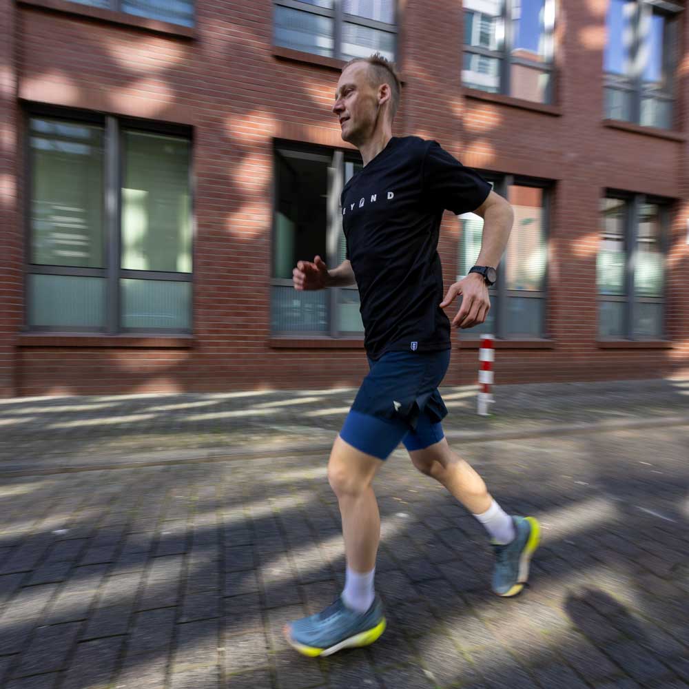 Ein fröhlicher Läufer in sportlicher Kleidung joggt energisch auf einem urbanen Gehweg, umgeben von modernen Gebäuden.